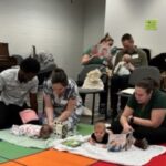 Parents try out Tummy Timmy by putting their babies on their bellies and opening up a cardboard book in front of them at a Montgomery County Library Hatchlings' session.